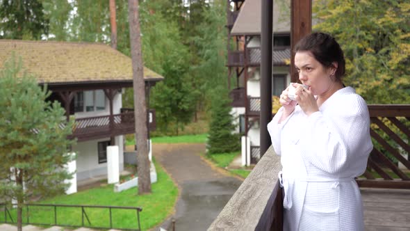 A Girl in a White Coat Goes Out on Balcony in Morning to Drink Coffee in Fresh Air