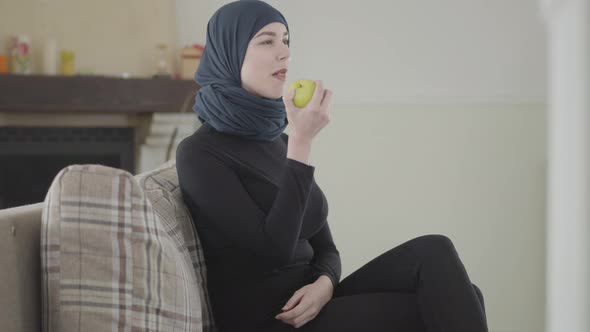 Beautiful Muslim Woman Watching TV Show and Biting Apple Wearing Traditional Headscarf on the