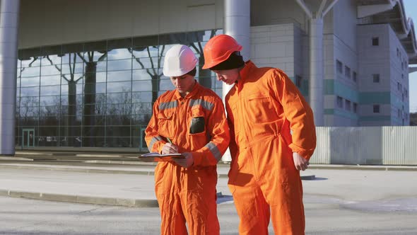 Two Construction Workers in Orange Uniform and Helmets Looking Over Plans Together