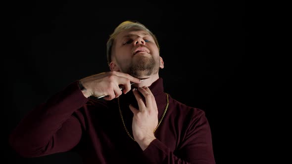 Portrait of Unshaven Young Man in Trendy Stylish Clothes Shaving with Knife Against Black Background