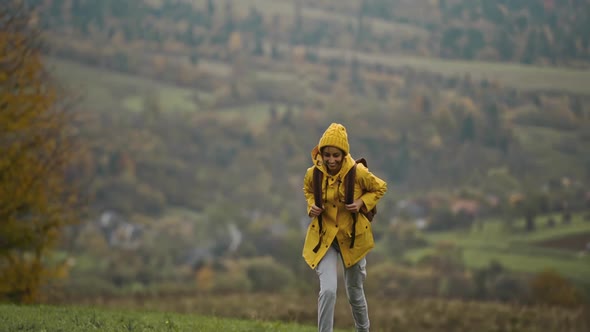 Beautiful Woman in Yellow Raincoat and Backpack Stands Against Fields and Mountains Raises Her Arms