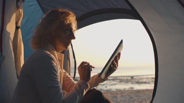 Side View of Mature Woman Lit By Sunset Light Relaxing in Camping Tent Using Tablet Pc