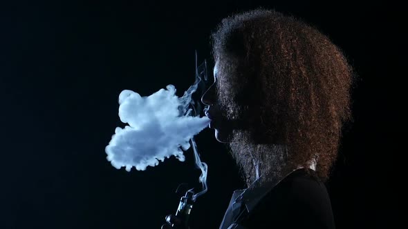 African American Woman Smokes an E-cigarette and Lets Smoke Out of Her Mouth. Black Background.