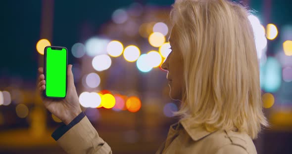 Bokeh Shot of Senior Woman Holding Smartphone with Blank Green Screen Outdoors