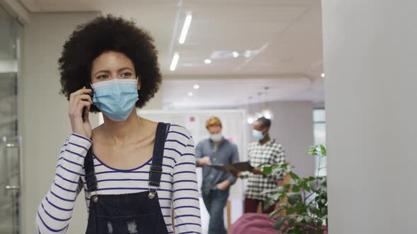 Portrait of african american creative businesswoman wearing face mask using smartphone in office