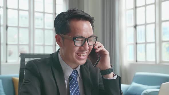 Asian Businessman Answering The Phone Call While Using The Computer For Working At Home