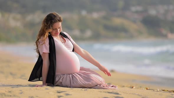 Pregnant Woman Sitting on the Beach Wearing Pink Dress and Black Vest