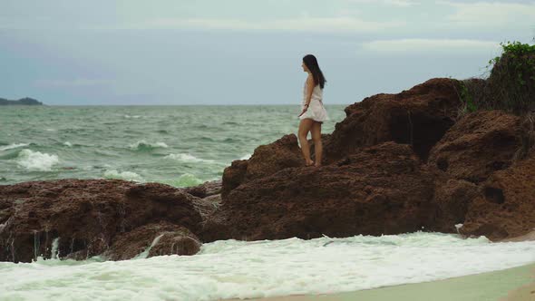 Young brunette woman staying on rocks near sea on stormy weather. Big waves hitting rocks while youn