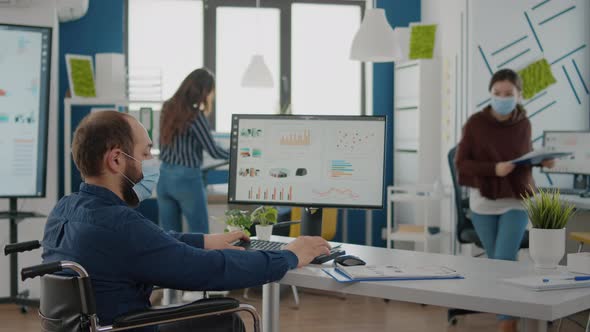 Invalid Worker in Wheelchair Talking with Colleague Typing on Computer