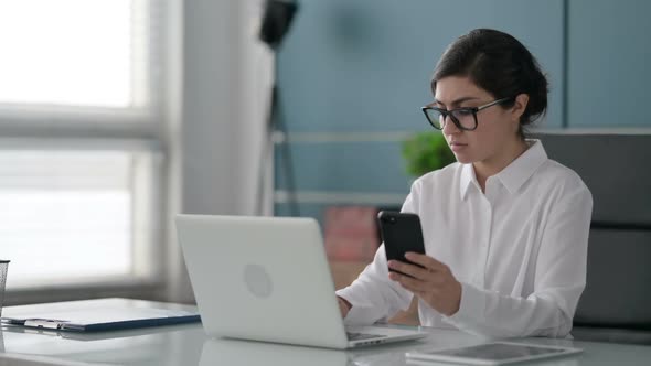 Indian Businesswoman using Smartphone while using Laptop in Office