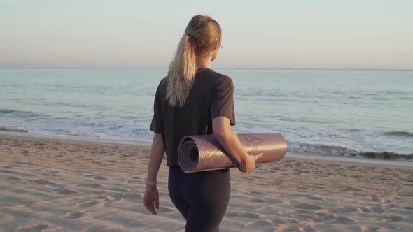 Travel girl walking towards calming ocean on beach with a yoga mat.