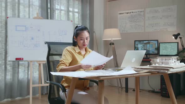 Asian Woman Engineer With A Laptop Looking At Blueprint While Working At The Office