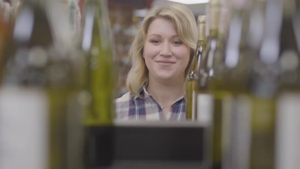 Face of Positive Caucasian Blond Woman Appearing Between Bottles of White Wine, Smiling Lady Taking