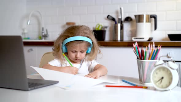 Little Girl in Headphones and in Front of a Laptop at Home in the Kitchen Draws Learning Online