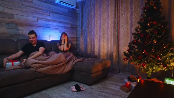 Young Couple Sitting on a Couch Under a Blanket  the Man Gives His Girlfriend a Christmas Gift