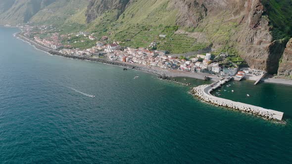 Seaside town below coastal cliffs, boat cruising to harbor, Madeira; drone