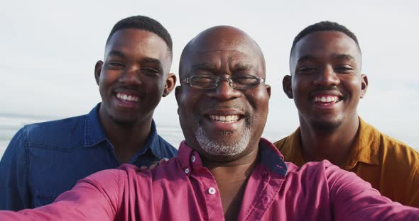 Smiling african american senior father and twin teenage sons standing on a beach taking a selfie