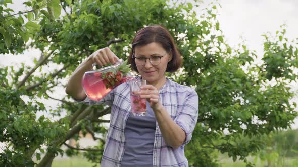 Middle Aged Woman Pouring Natural Drink with Strawberries and Mint From Jug Into Glass