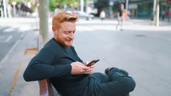 Relaxed ginger man texting by smartphone while sitting on the bench