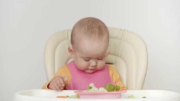 A newborn child aged one year is sitting on a feeding chair and eating a fresh green cucumber.