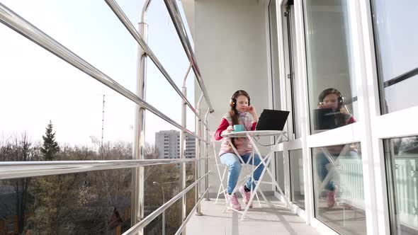Teenager Girl in Headphones, Working on Laptop, Chatting Online, on Open Balcony. Spring Sunny Day
