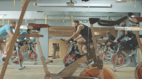 Joyful Professional Trainer Endorsing People Riding Fast on Exercise Bikes in Gym. Wide Shot Side