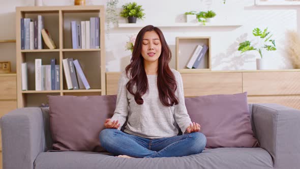 Clam of Asian young woman doing yoga lotus pose to meditation and relax on couch during work online