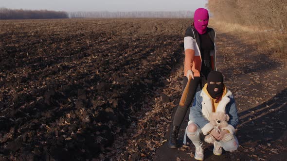 Women in Balaclava with Baseball Bat and Teddy Bear in Countryside