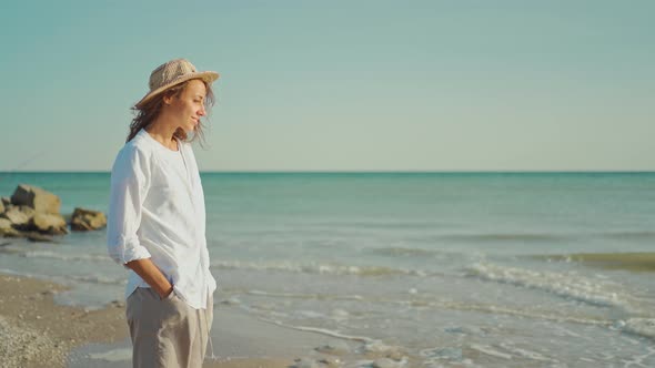 Attractive Smiling Woman Traveler in White Shirt and Straw Hat Walking Along Sea Coastline