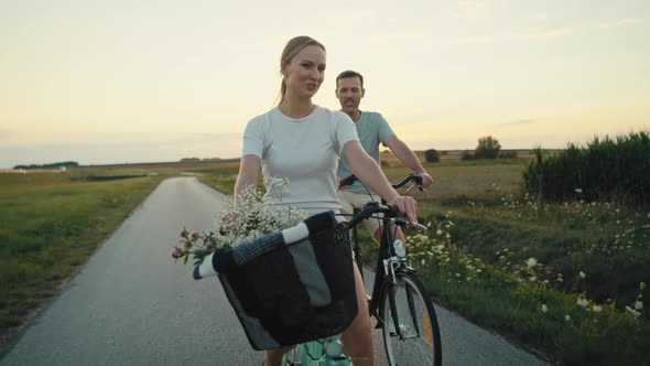 Front view of cheerful caucasian couple of middle age riding a bike on sunset on village road. Shot