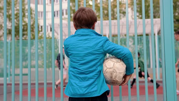 A Little Insecure Boy Holding Deflated Ball and Watching Other Kids Playing Football