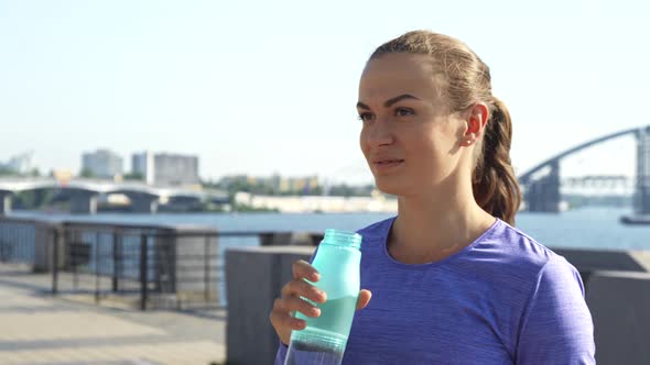 Woman Drinks Water and Shows Thumb Up Against Urban Background