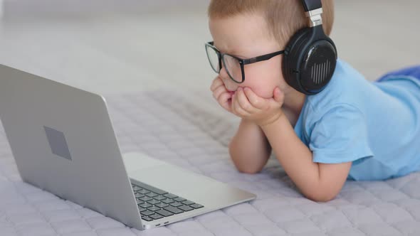 Baby Child Boy in Blue Clothes Lying on Floor in Headphones Watching Laptop