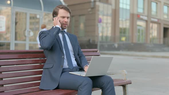 Businessman Talking on Phone and using Laptop while Sitting Outdoor on Bench