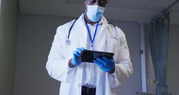 African american male doctor wearing face mask using digital tablet in hospital patient room