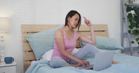 Happy asian woman sitting on bed, using laptop in the morning