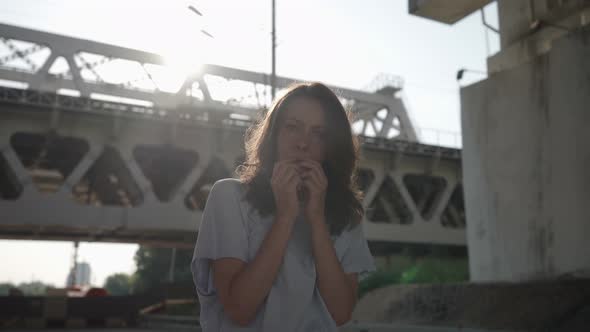 the Brunette Stands and Covers Face with Hands Against the Background of the Railway Bridge
