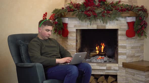 A Young Man Sits in an Armchair By the Fireplace with Horns on His Head and is Typing on a Laptop