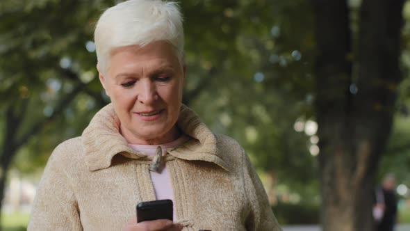 Elderly Woman of Retirement Age with Short Gray Hair Walks in Park Outdoors Holding Phone