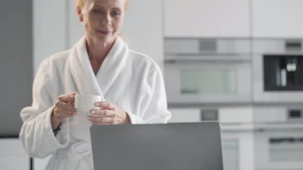 Senior Woman in Bathrobe Working Remotely on Laptop From Home Drinking Coffee in Kitchen