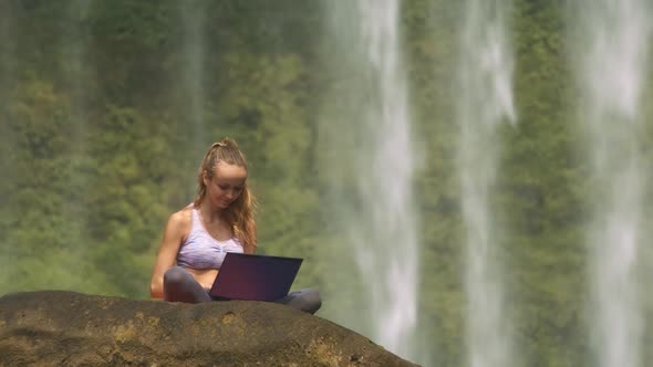 Girl Puts Notebook on Stone and Types at Waterfall