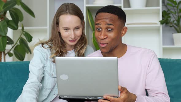 Cheerful Interracial Couple Web Surfing on Laptop Together, Sitting on Sofa at Home