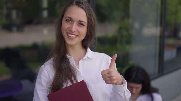 Positive Woman with Laptop Gesturing Thumb Up Smiling Looking at Camera Standing on City Street with