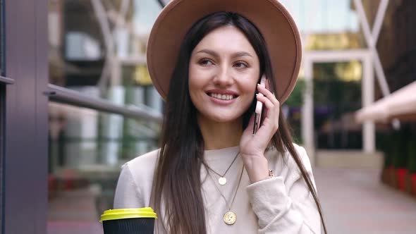 Smiling Happy Woman with Stylish Hat Walking Near Glass Wall and Enjoying Her Phone