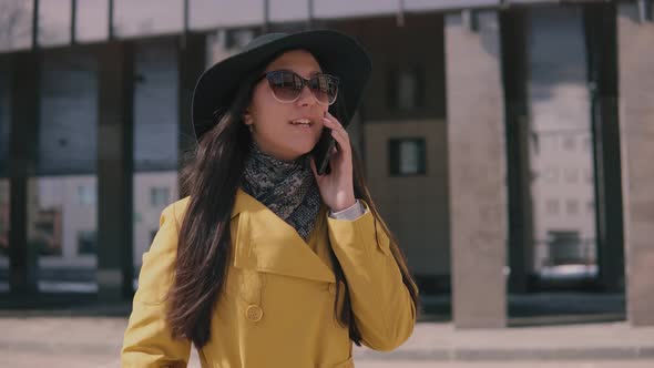 Stylish Young Girl in a Yellow Raincoat Hat and Glasses Talking on the Phone and Laughs