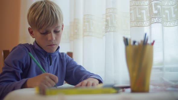 The student gets a pencil from a glass and begins to draw. A child draws near a window at a table.