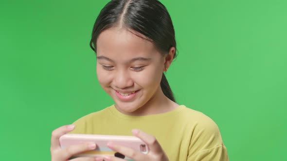 Smiling Young Asian Kid Girl Looking At The Phone Screen On Green Screen In The Studio