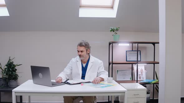 Male Doctor Talking and Waving in Video Chat on Laptop in White Office