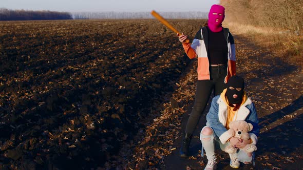 Women in balaclava with baseball bat and teddy bear in countryside.