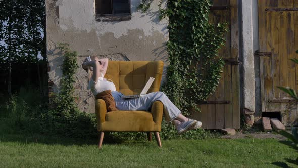 Woman sitting in an armchair using laptop in a garden with wall and doors on background